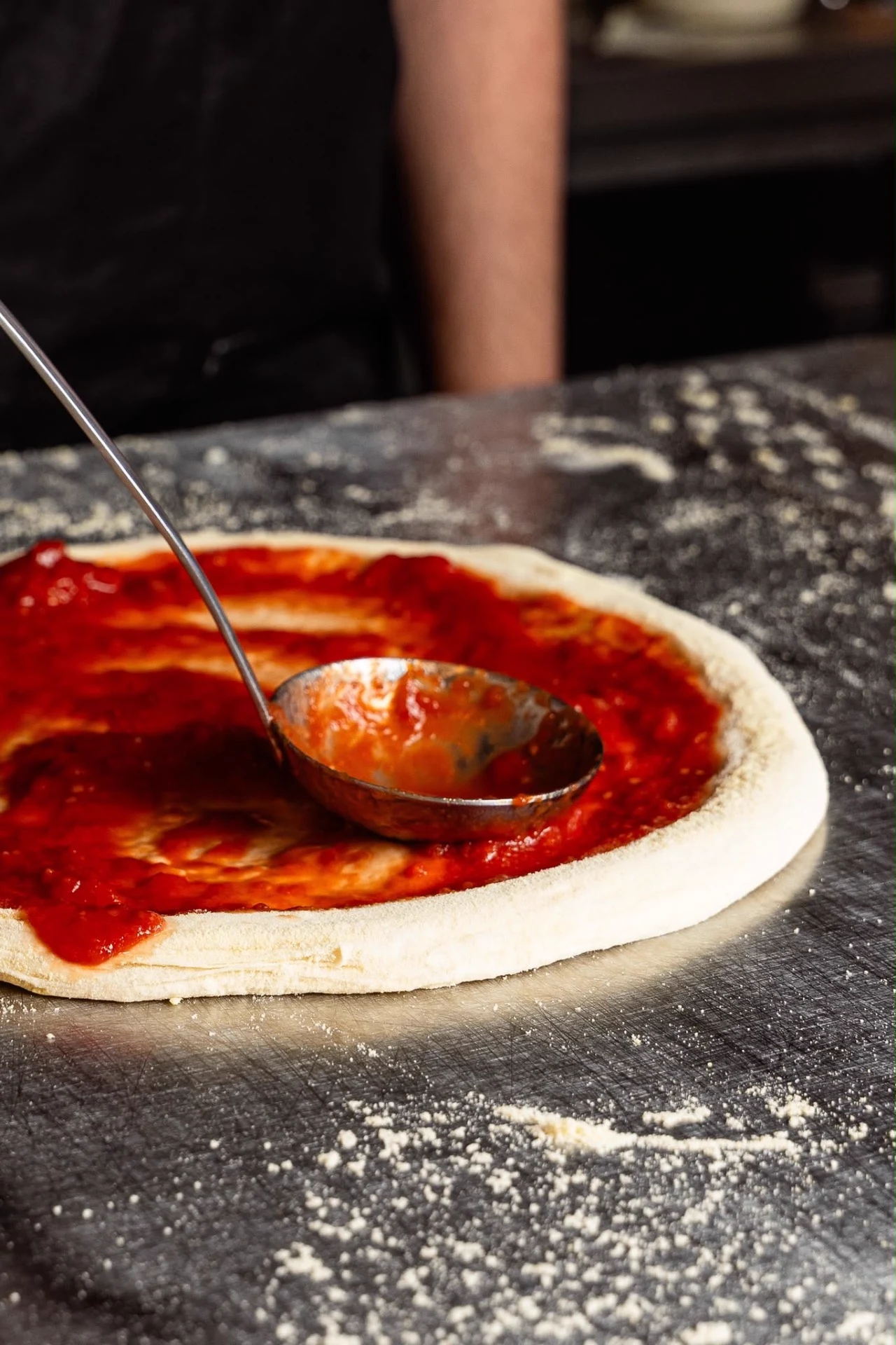 Molho de tomate a ser espalhado sobre a massa crua numa bancada polvilhada de farinha